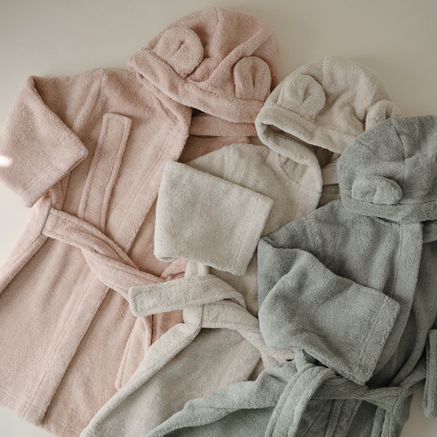 Set of pink, beige, and gray bathrobes with matching hats on a neutral background
