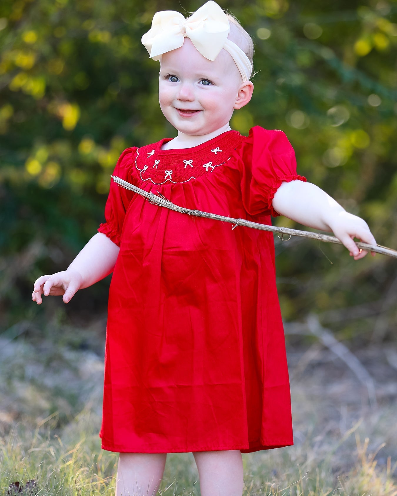 Child in a red dress holding a stick outdoors with greenery in the background
