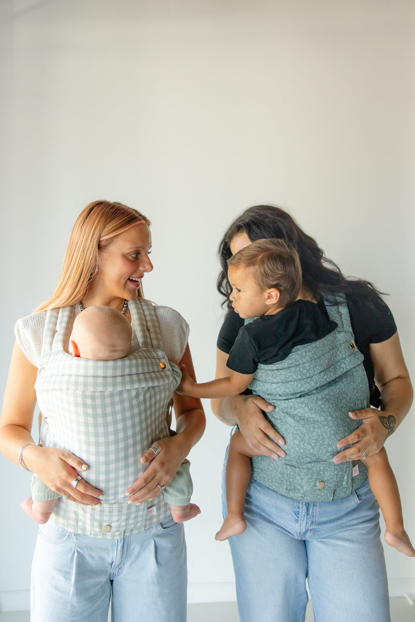 Two women holding babies in baby carriers against a white background
