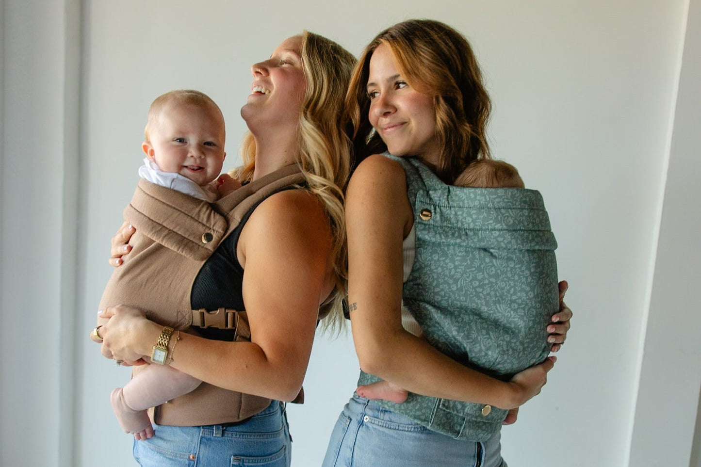 Two women holding babies in baby carriers against a plain background