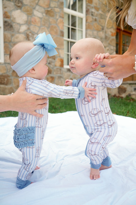 Two babies in matching outfits standing on a blanket outdoors.