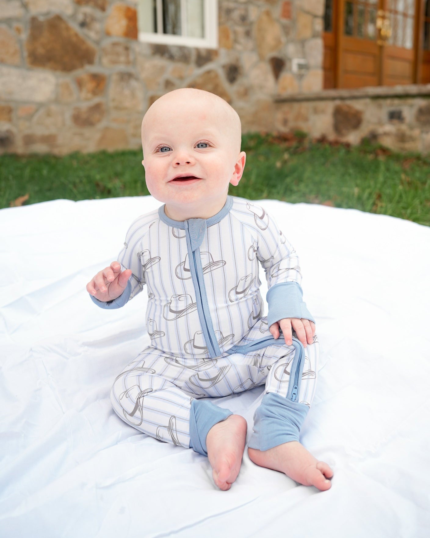 Baby in a patterned onesie sitting on a blanket outdoors with a stone building in the background