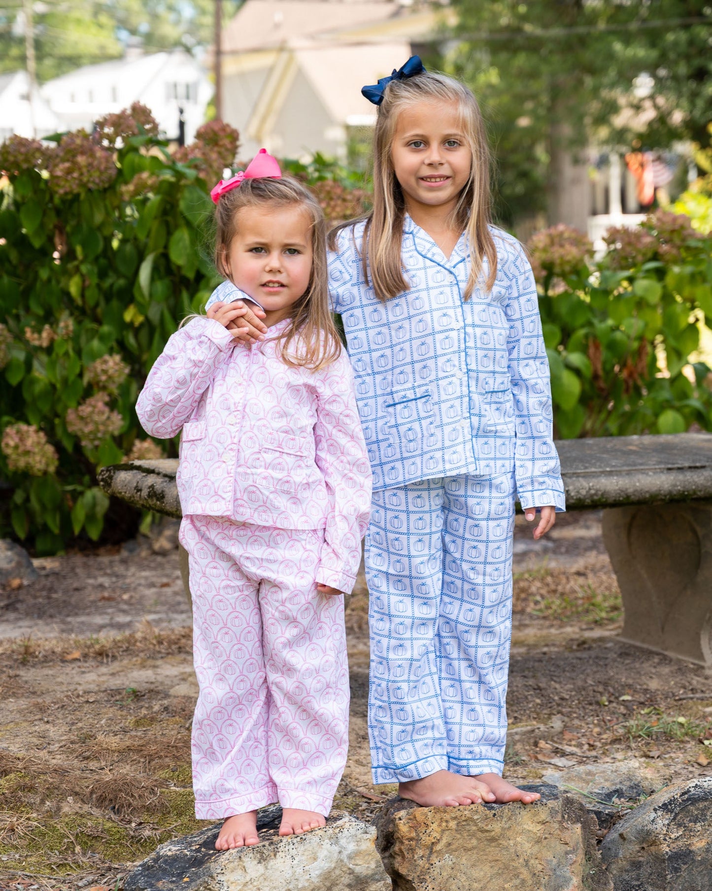 Two young girls in pajamas standing outdoors on a stone surface with greenery in the background.