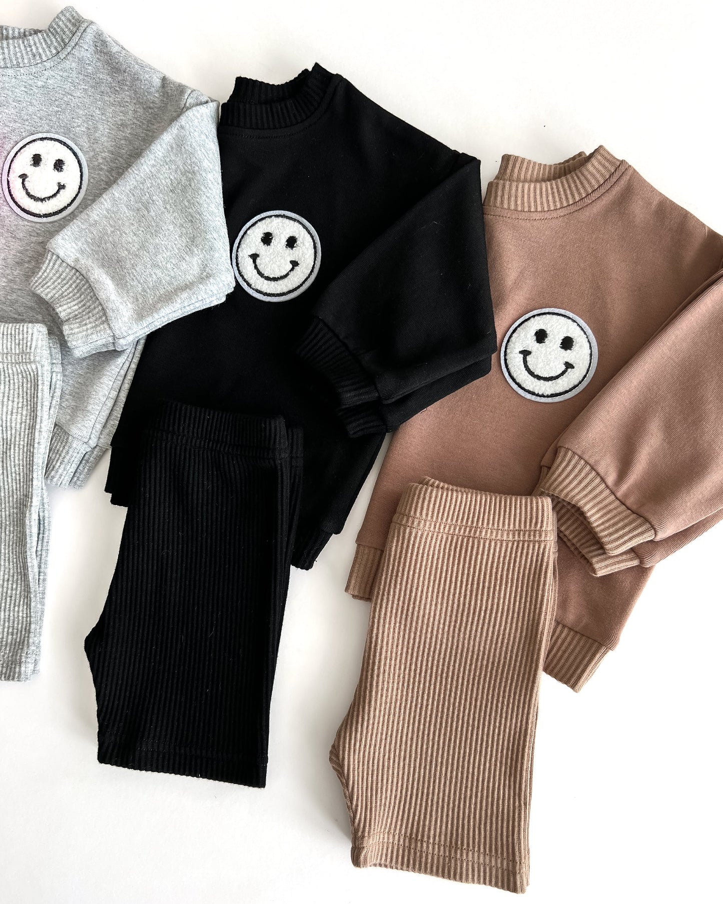 Three sweaters in gray, black, and brown with smiley face designs on a white background