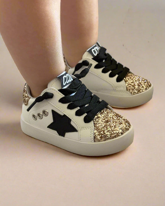 White sneakers with gold glitter and black stars worn by a toddler on a carpeted floor.