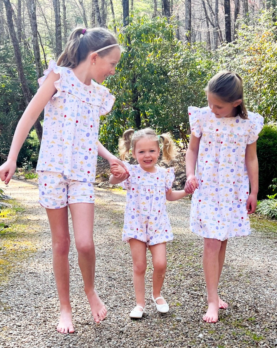 Three young girls wearing matching dresses standing outdoors on a gravel path with trees in the background.