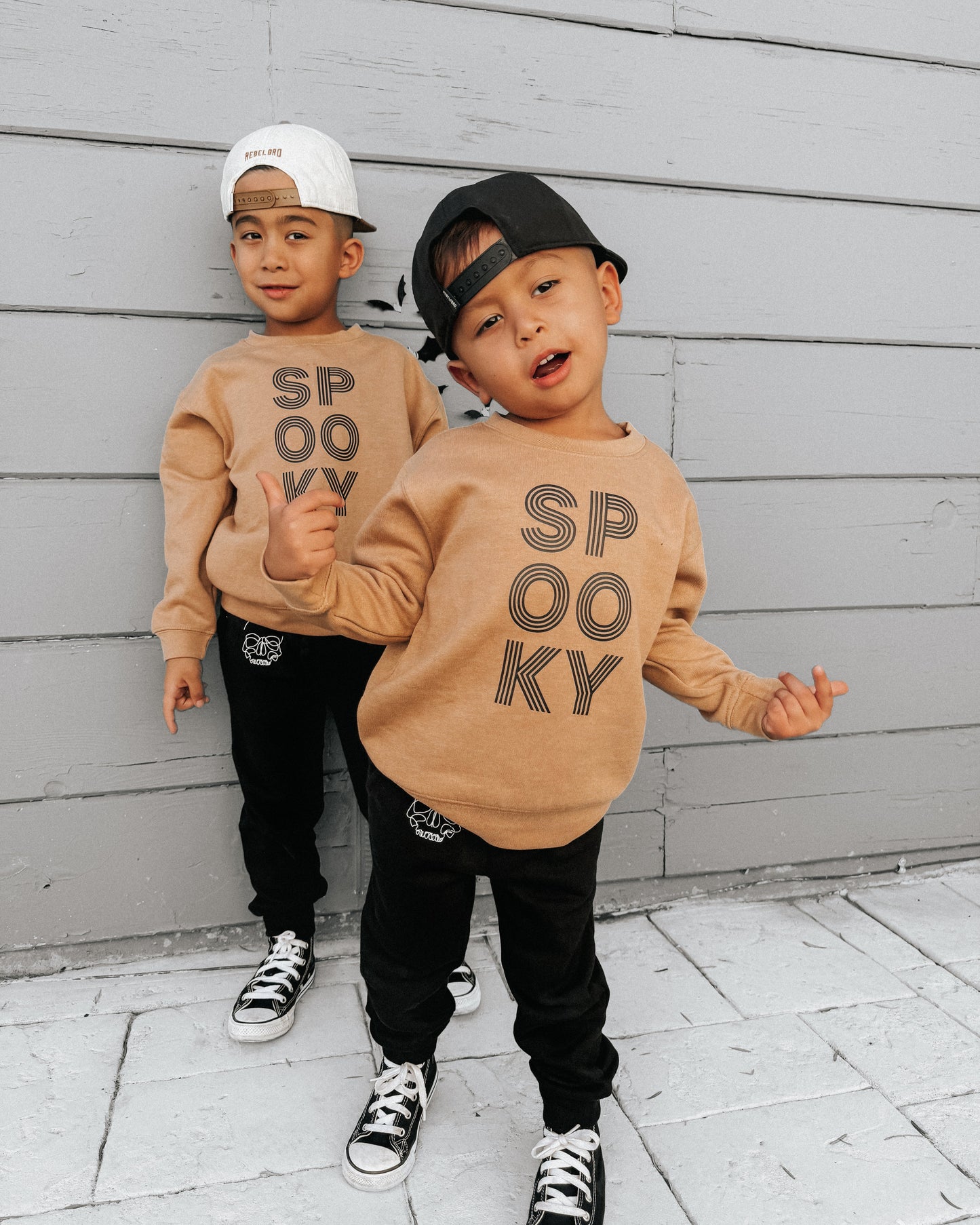 Two children wearing beige sweatshirts with 'SPooky' text, standing against a gray wooden wall.
