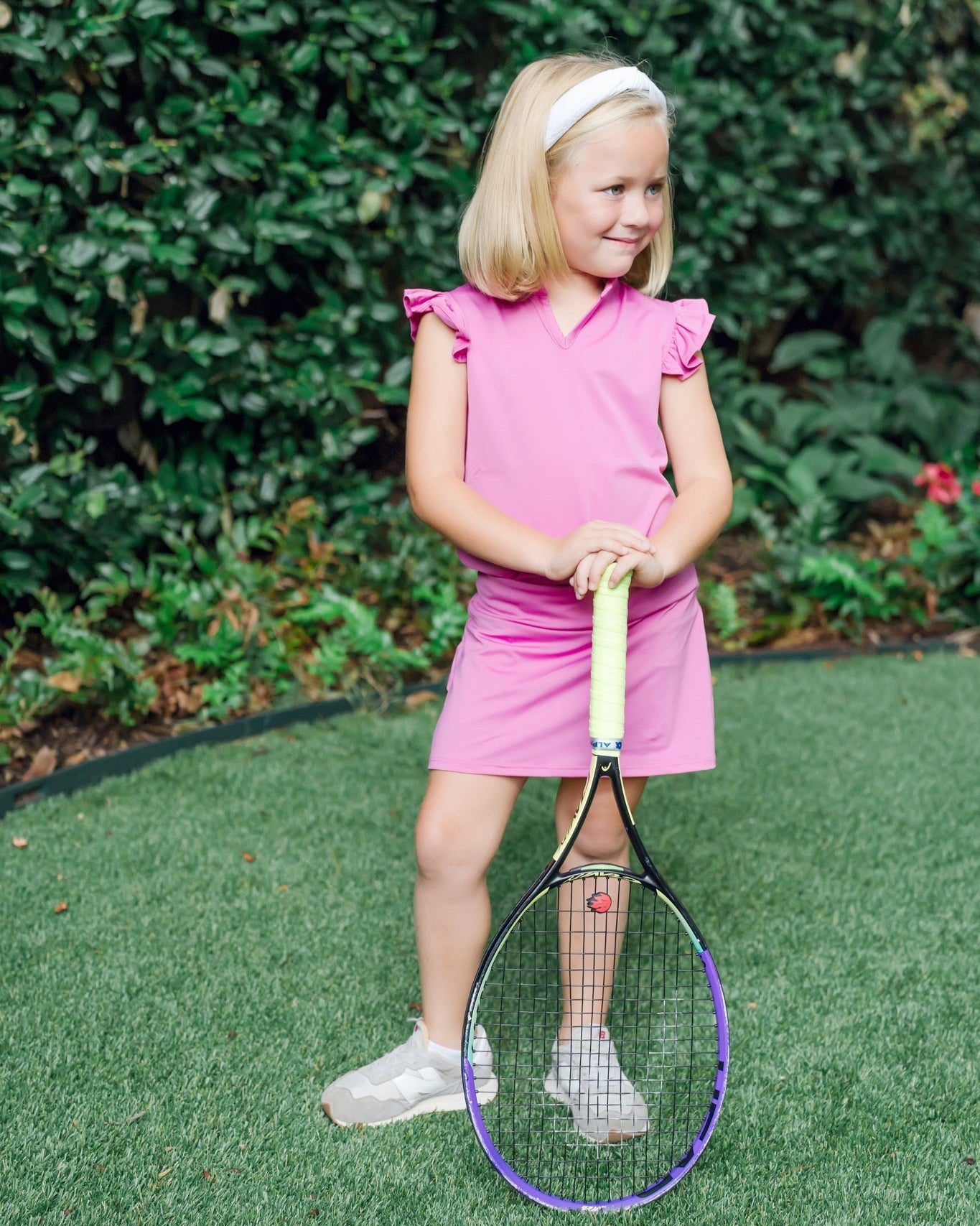 Young girl in a pink set holding a tennis racket on a grassy area with greenery in the background