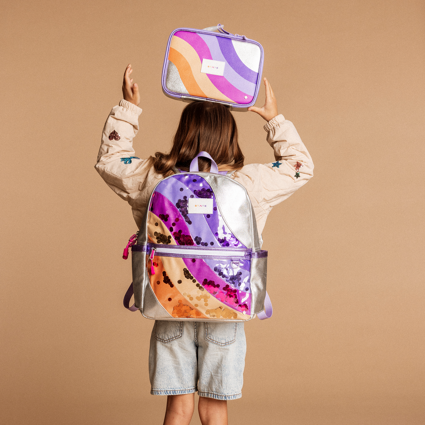 Person holding a colorful backpack and lunch box against a beige background
