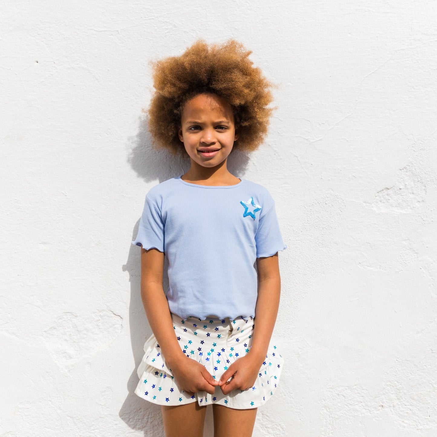 Young girl wearing a light blue t-shirt with a star design and a white skirt with blue patterns against a white wall.
