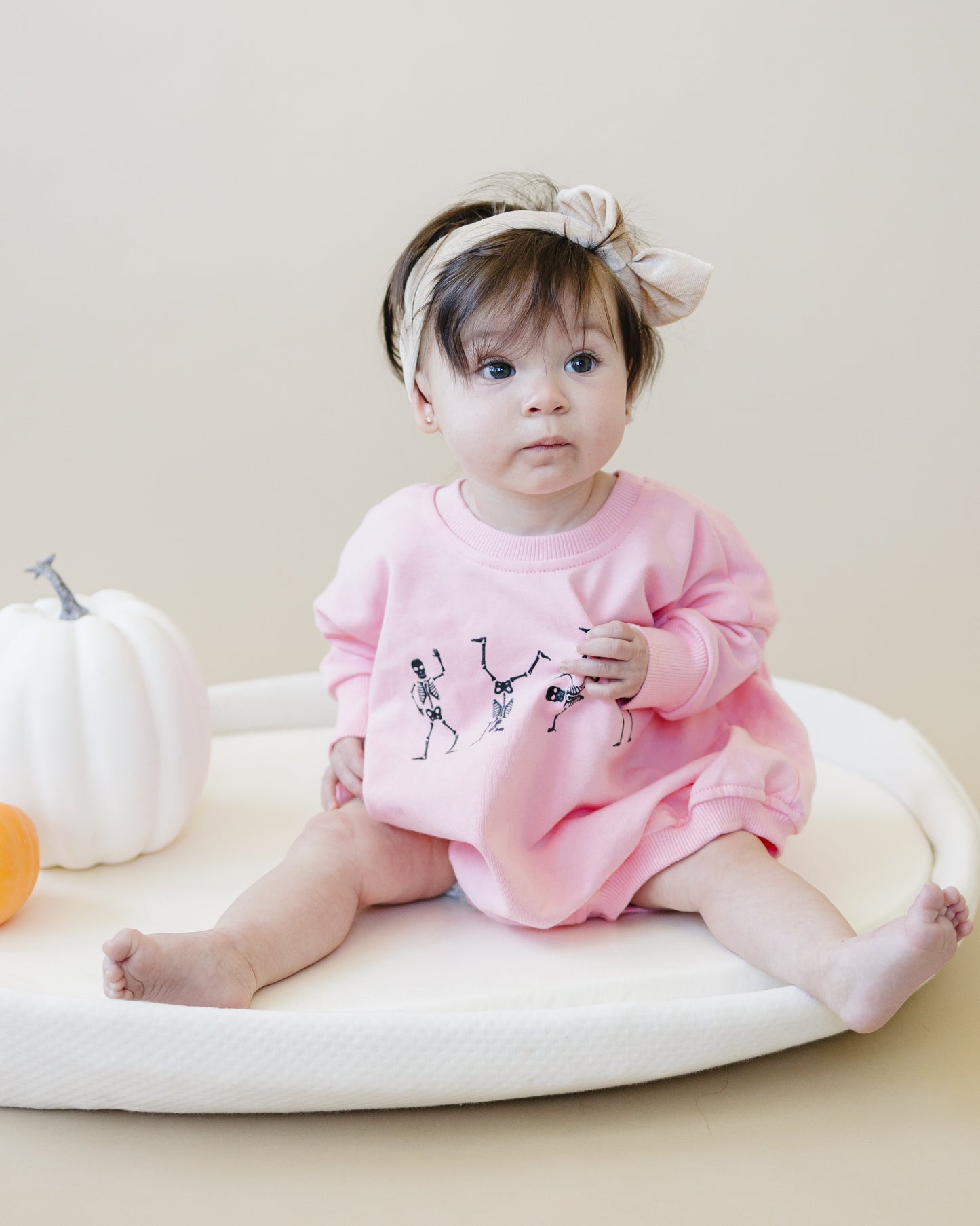 Baby in a pink outfit sitting on a white cushion with pumpkins around