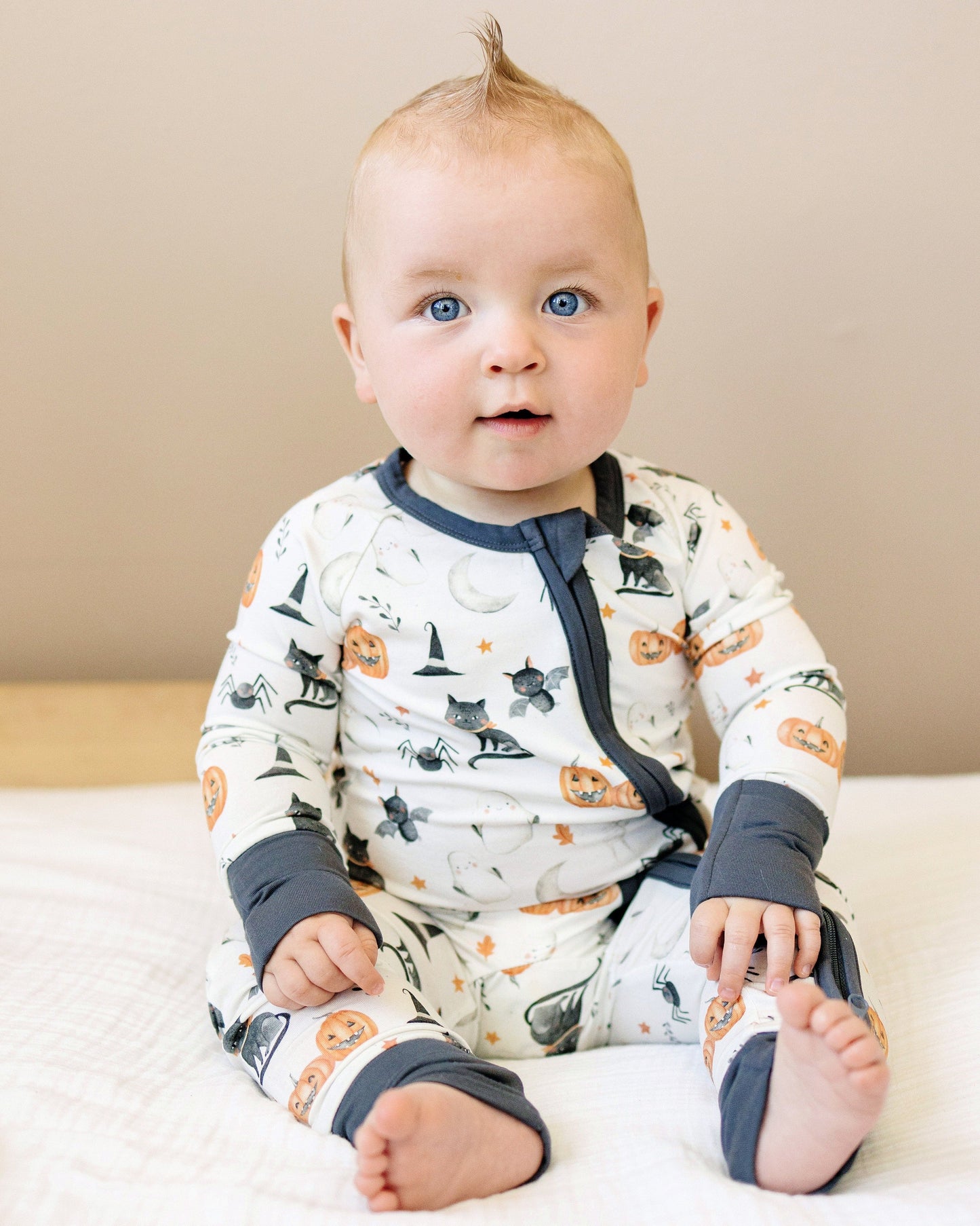 Baby wearing a Halloween-themed onesie sitting on a bed.