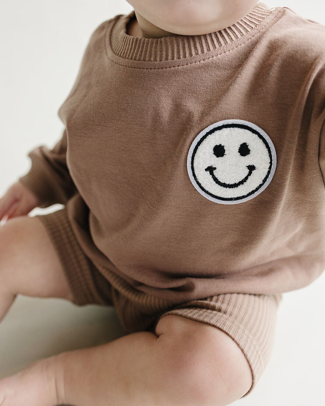 Child wearing a brown sweater with a smiley face patch on a plain background