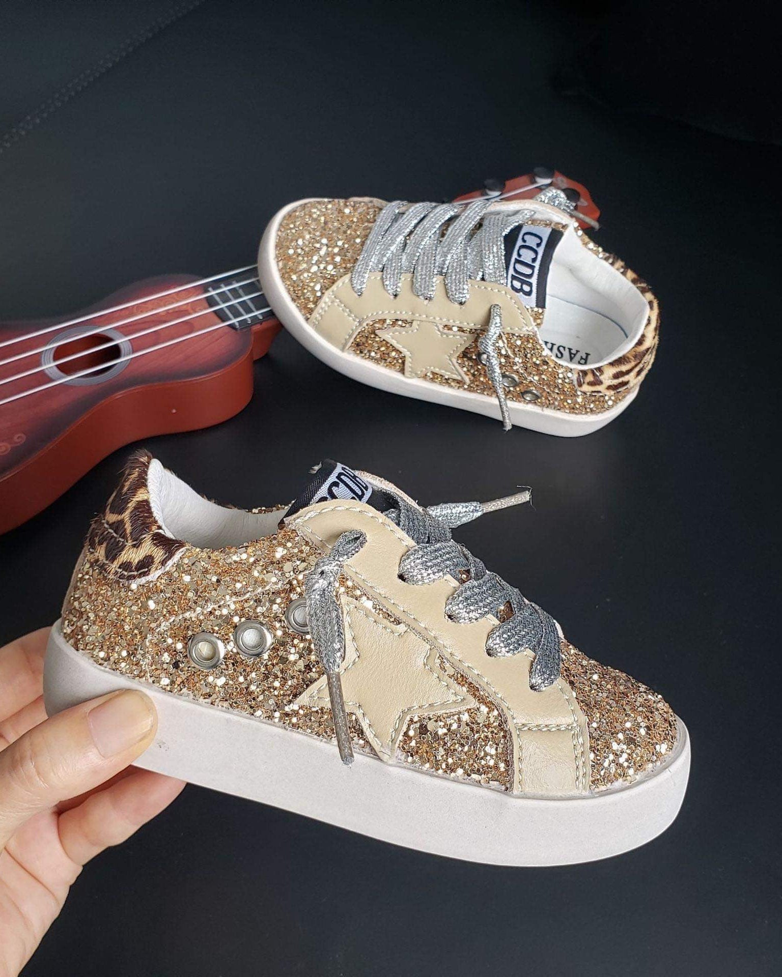 Sparkly sneaker with leopard print and pearl details held by a hand, with a small red guitar in the background.