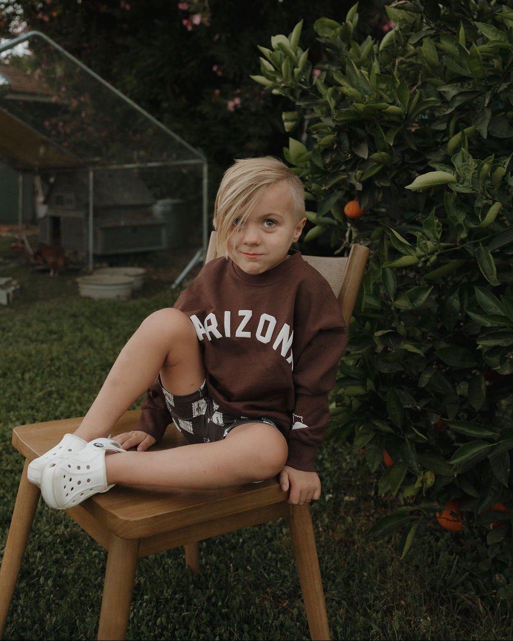 Child sitting on a wooden chair outdoors with greenery in the background