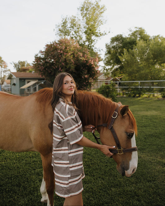 Woman standing next to a horse in an outdoor setting with trees and a house in the background.