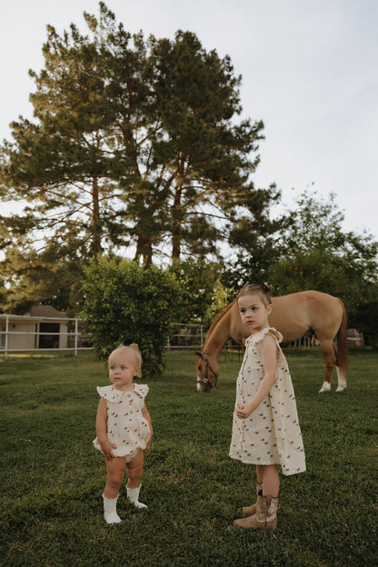 Two children in white outfits standing in a grassy field with a horse in the background.