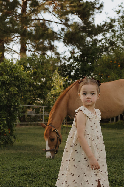 Young girl in a dress standing next to a horse in a grassy field with trees in the background
