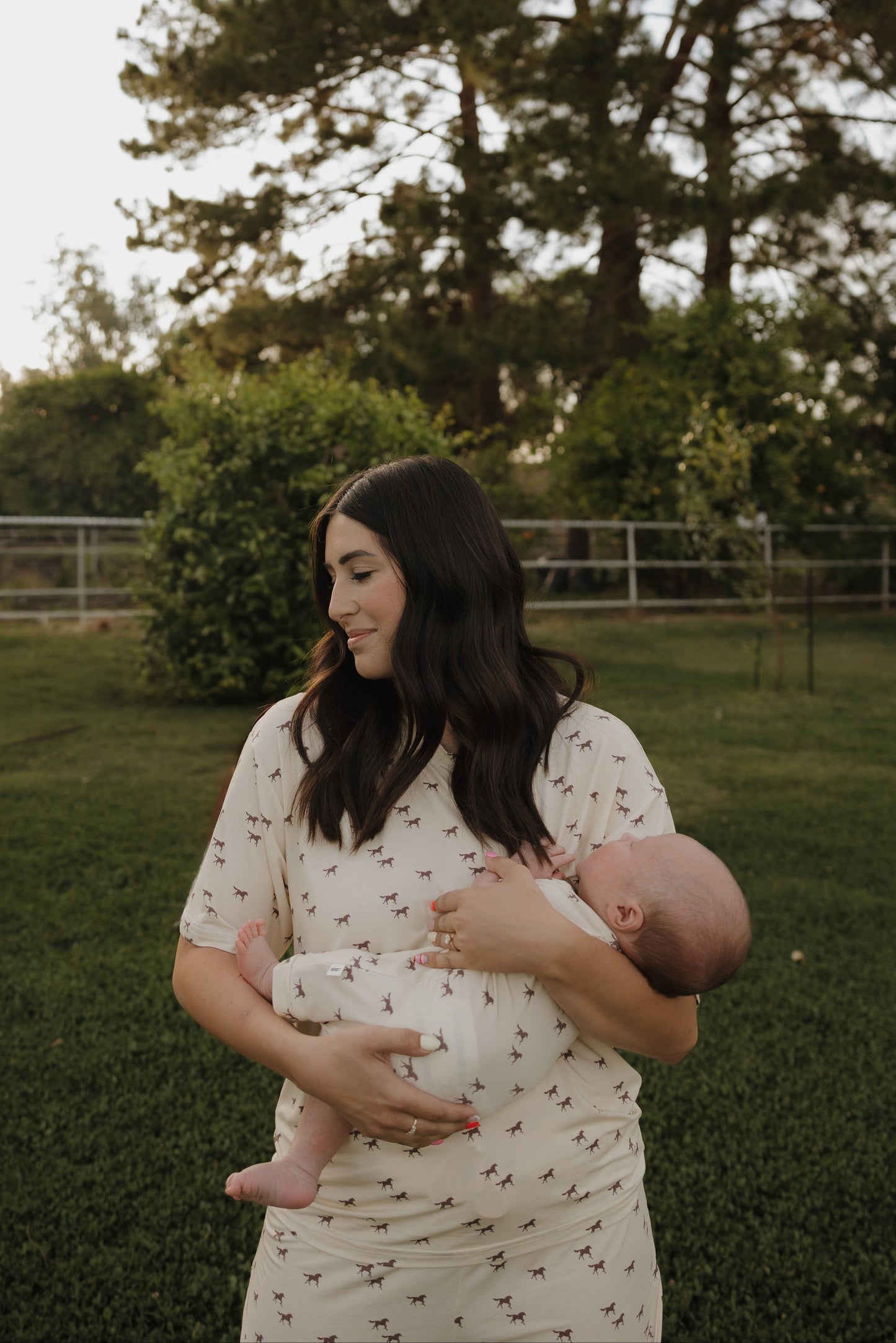 Woman holding a baby outdoors in a natural setting with trees and grass.