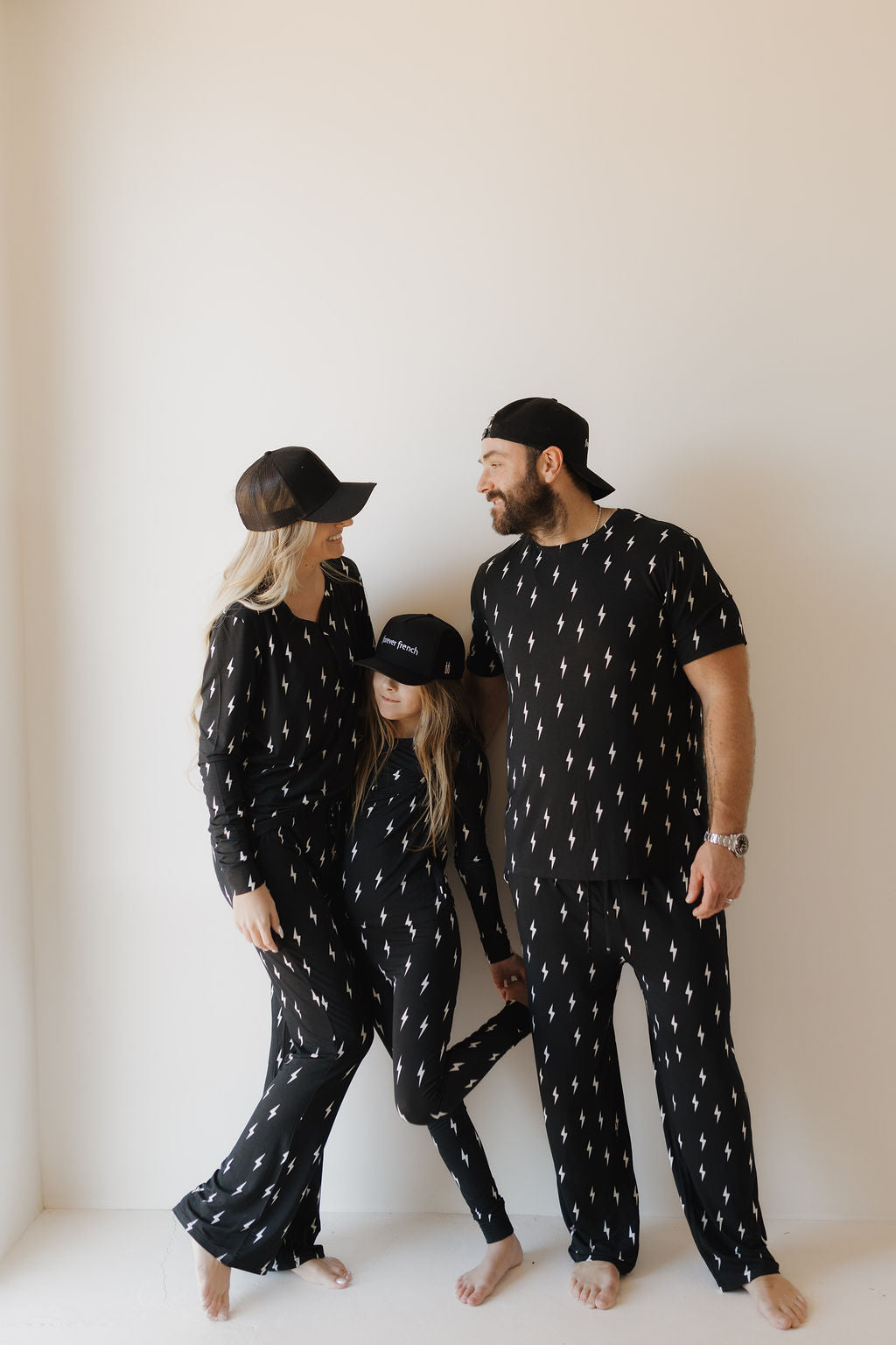 Family of three wearing matching black pajama sets with white patterns against a plain background