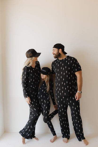 Family of three wearing matching black pajama sets with white patterns against a plain background