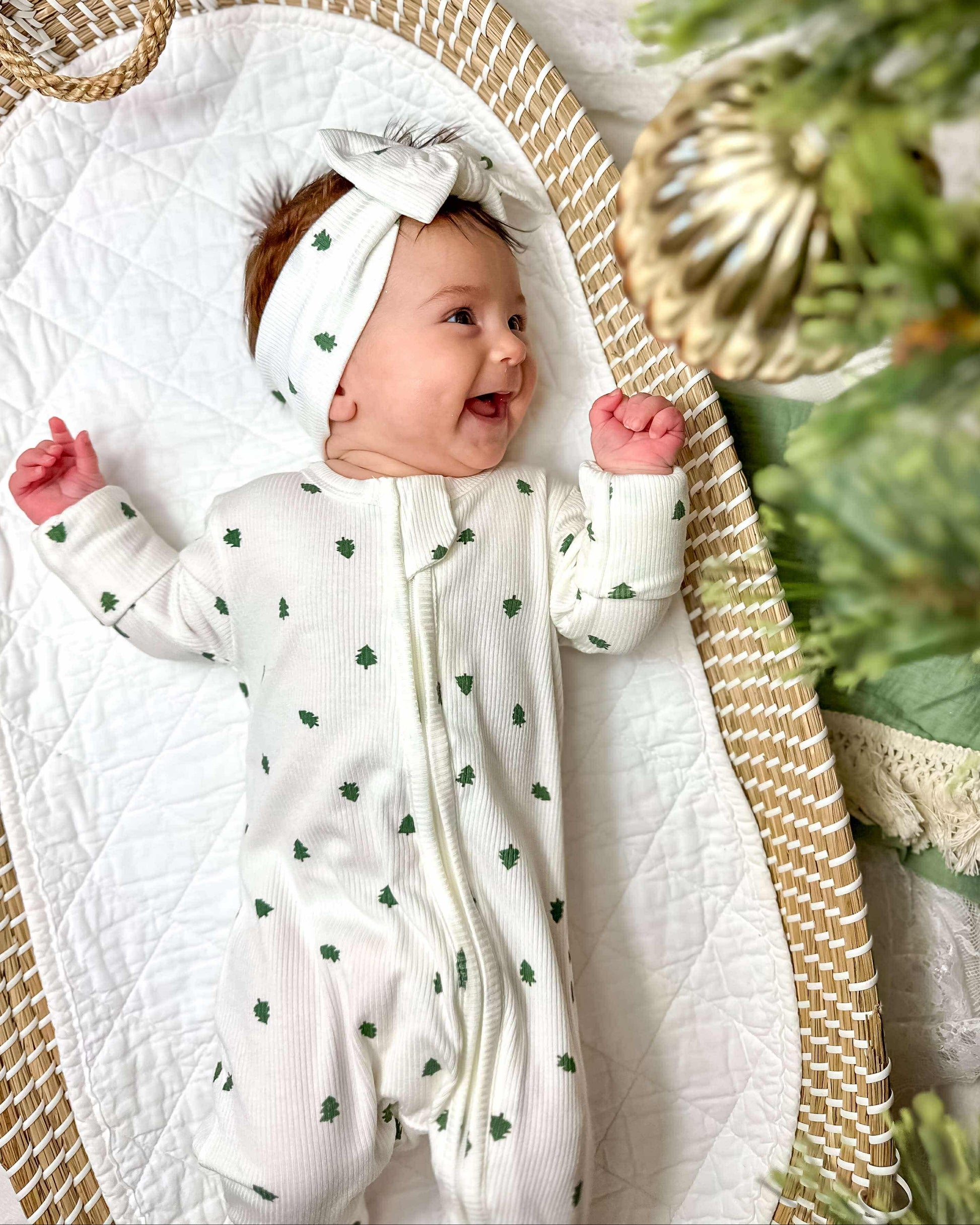 Baby in a white outfit with green patterns, wearing a matching headband, sitting in a wicker basket with a Christmas tree in the background.