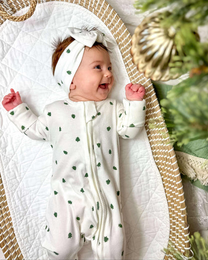 Baby in a white outfit with green patterns, wearing a matching headband, sitting in a wicker basket with a Christmas tree in the background.