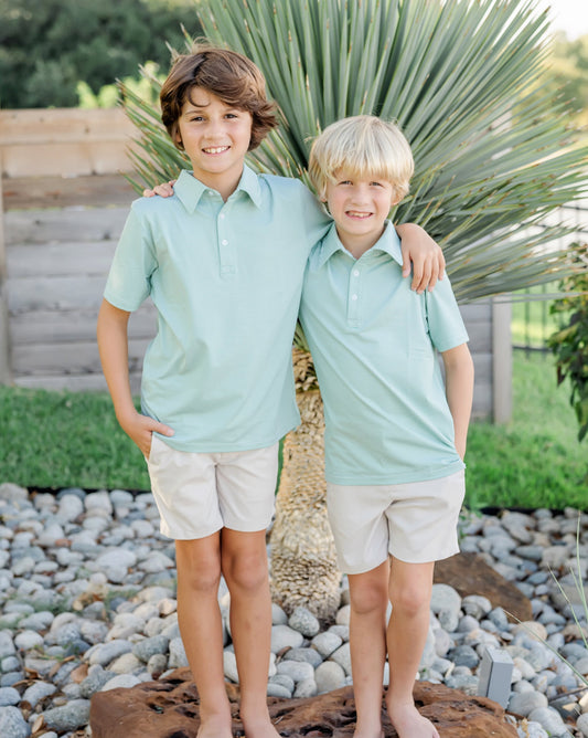 Two boys in matching in green shirts and beige shorts standing on a wooden log with a natural background.