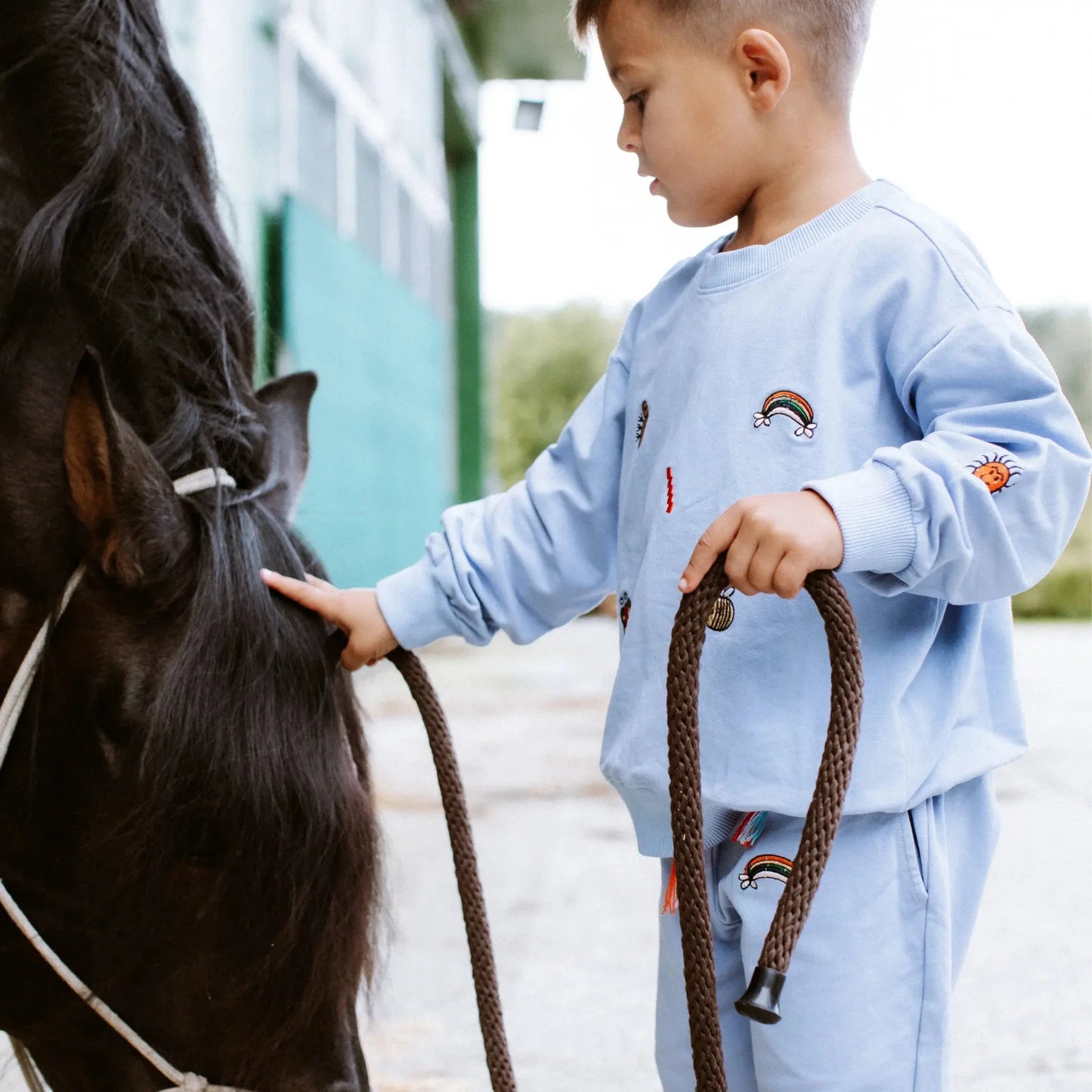 Child in a light blue outfit with rainbow designs petting a horse.