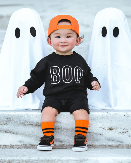 Child in Halloween costume with 'BOO' shirt and orange cap, standing between two ghost costumes.