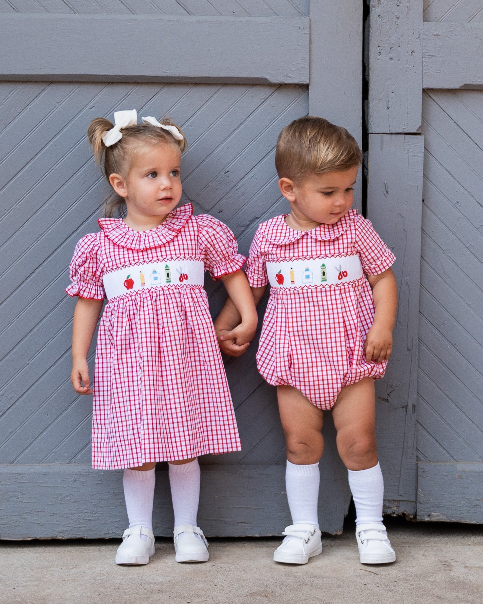 Two children in matching red and white checkered outfits standing in front of a gray wooden door.