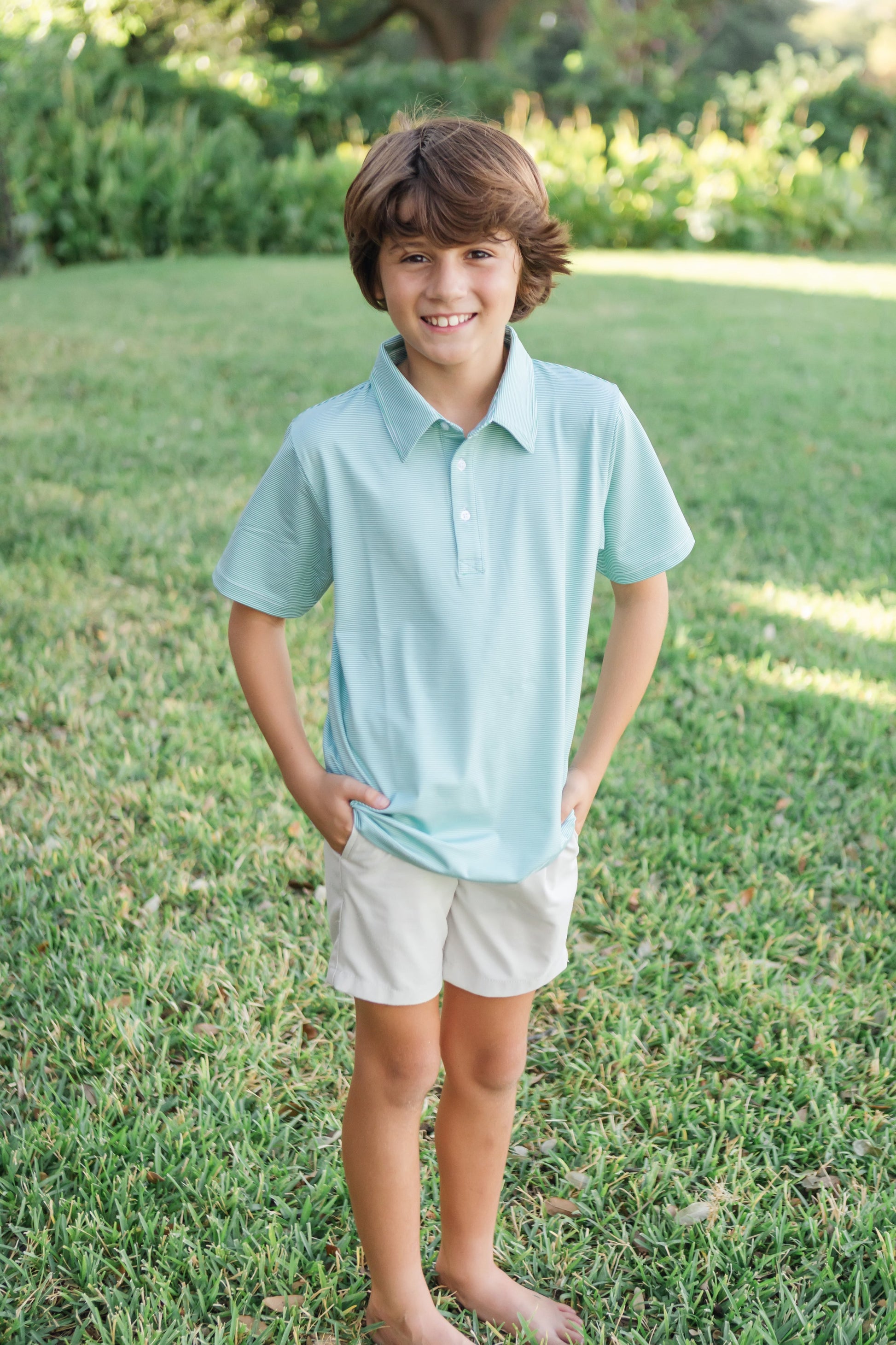 Young boy wearing a green striped polo shirt and white shorts standing on grass.