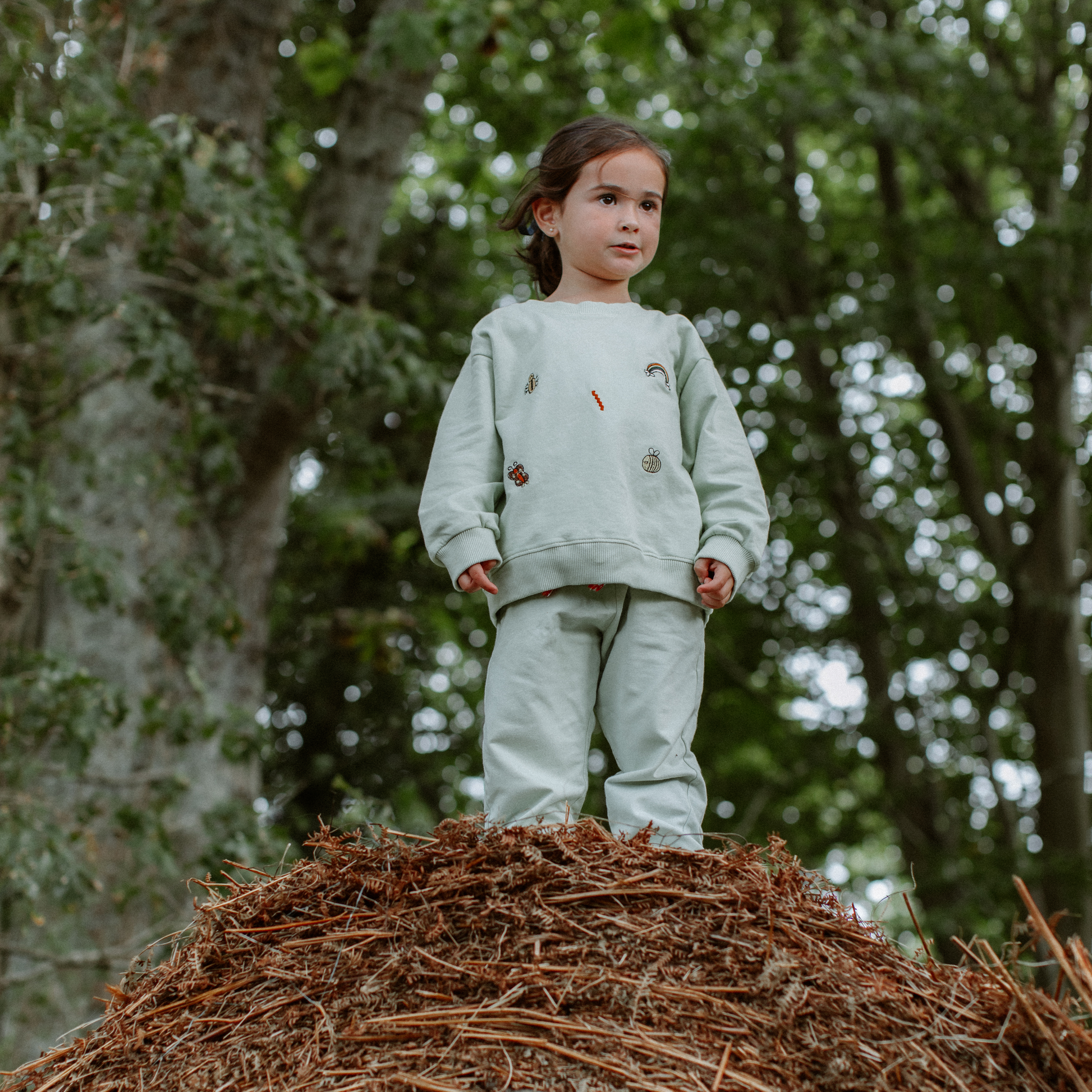 Child in light green outfit standing on a pile of wood chips with trees in the background