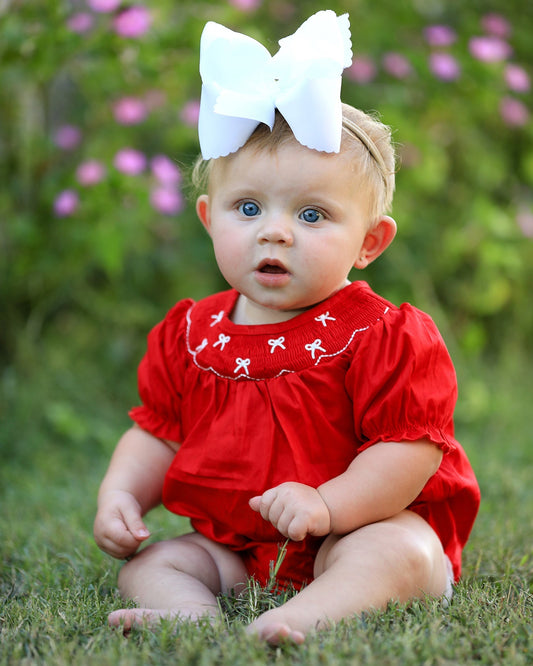 Baby in a red outfit with a large white bow sitting on grass.
