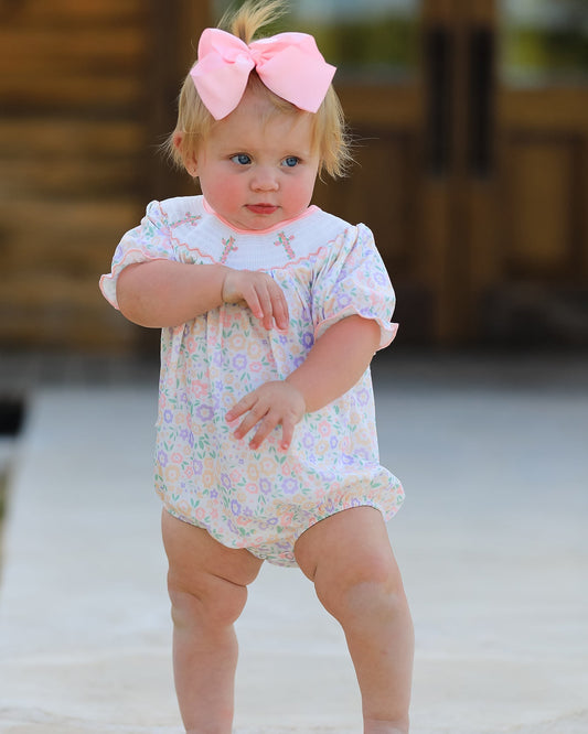 Baby in a floral outfit with a pink bow standing on a sandy surface.