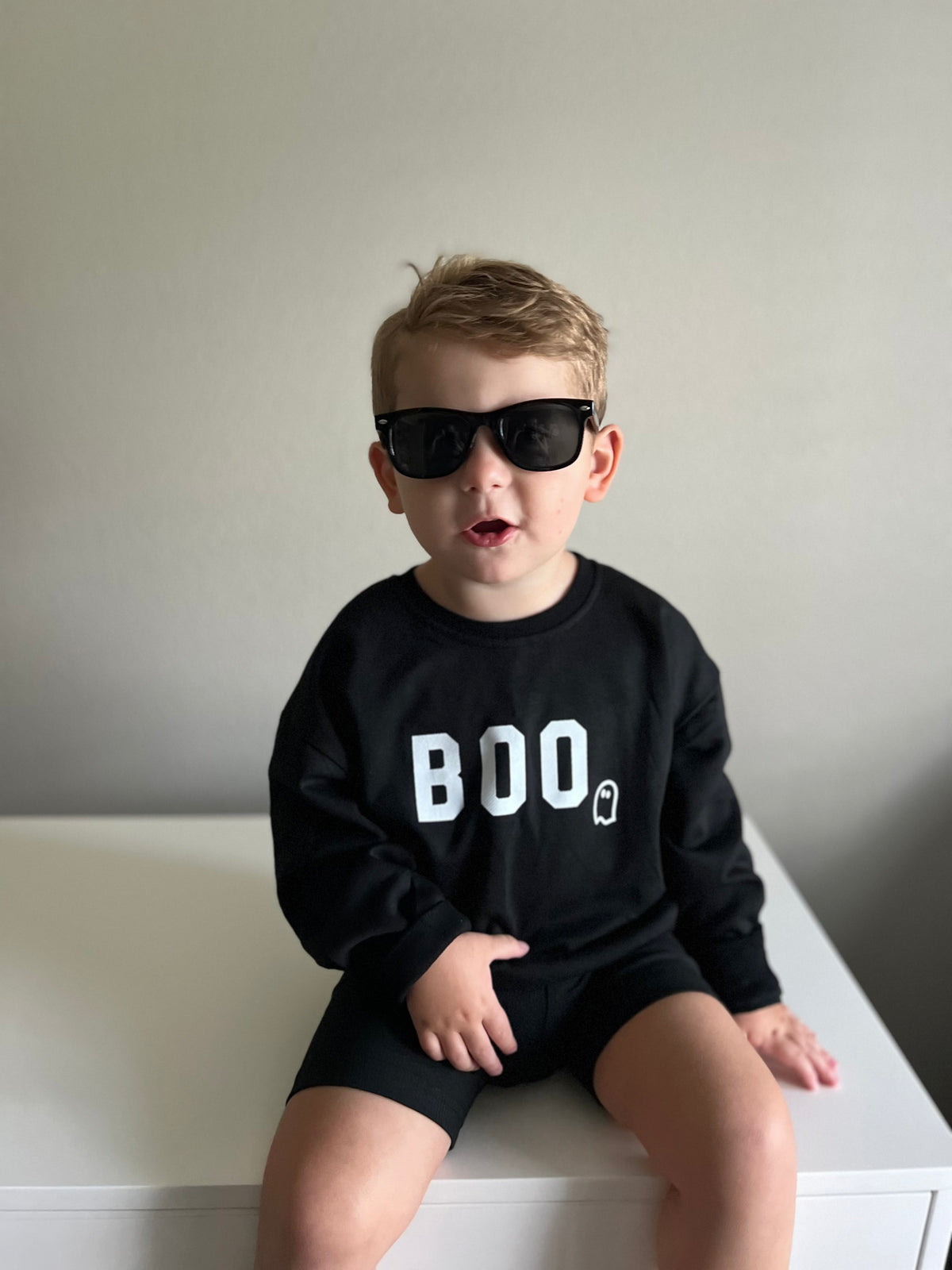 Child wearing a black 'BOO' sweatshirt and sunglasses on a white surface.