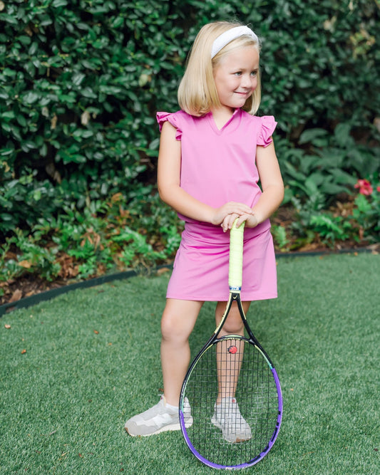 Young girl in a pink set holding a tennis racket on a grassy area with greenery in the background