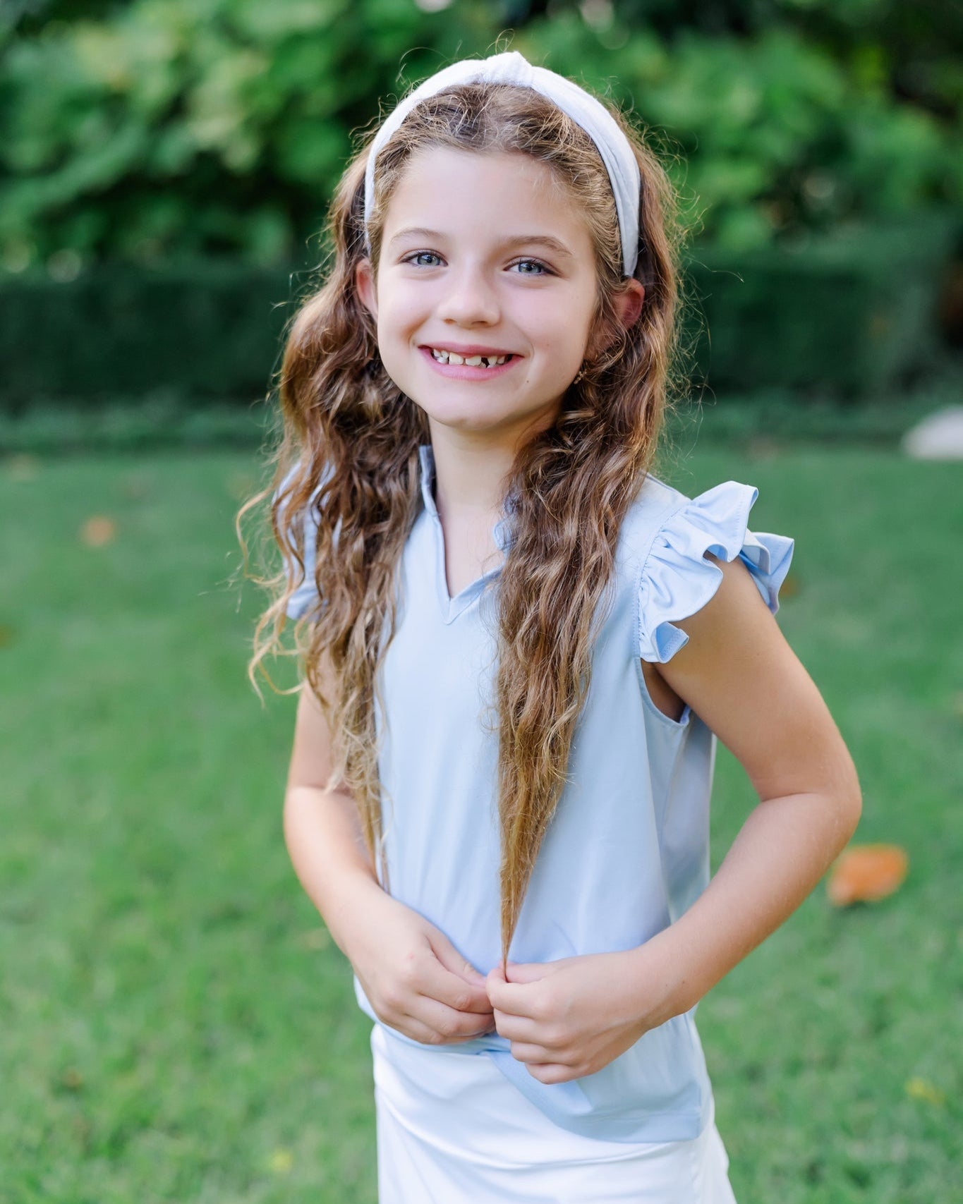 Young girl with long hair wearing a light blue sleeveless top with ruffled sleeves outdoors.
