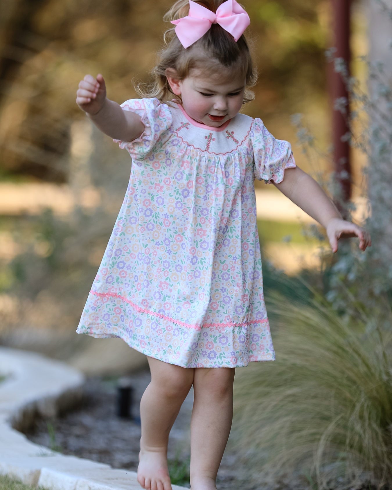 Young girl in a floral dress with a pink bow standing outdoors.