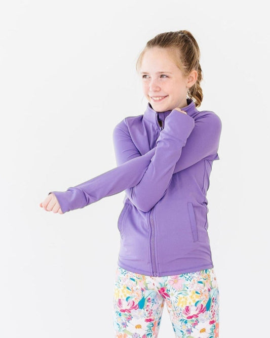Young girl in a purple jacket and colorful leggings standing next to a pink exercise ball and water bottle on a light background.