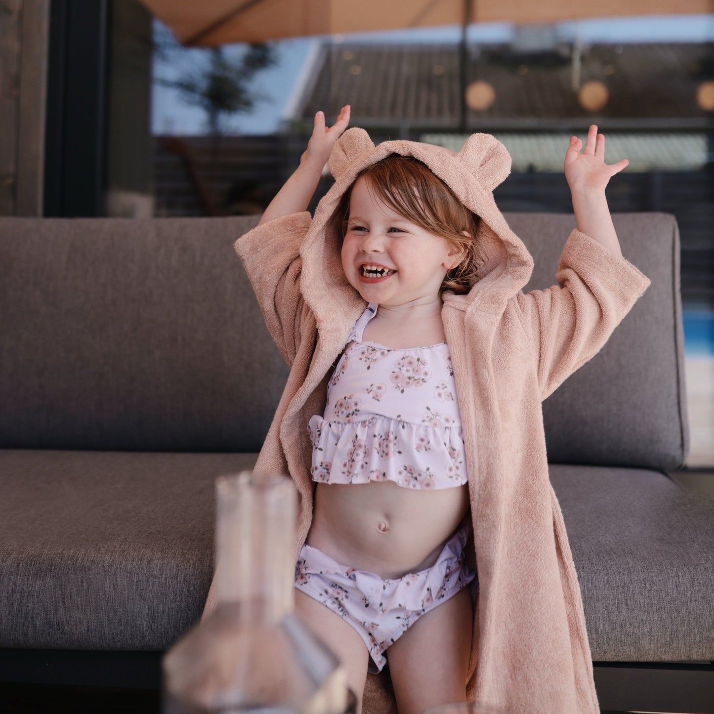 Child wearing a floral swimsuit and pink robe with bear ears, standing on a patio.