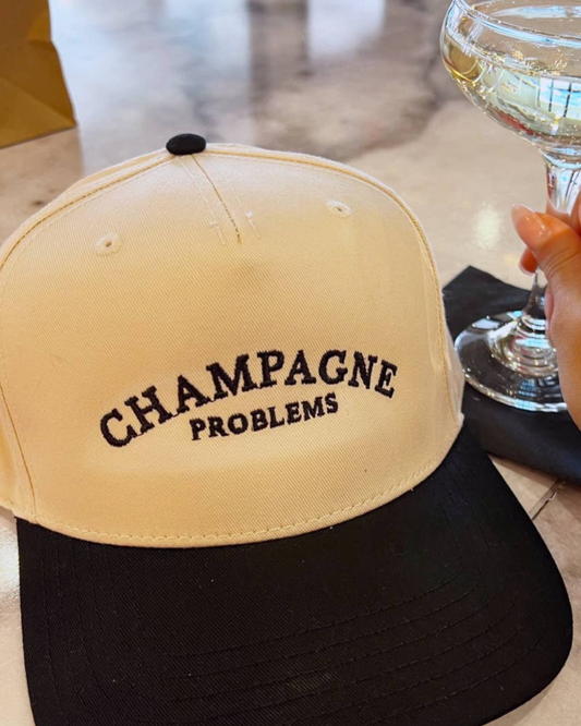 Beige cap with 'Champagne Problems' text and a hand holding a glass of champagne on a table.