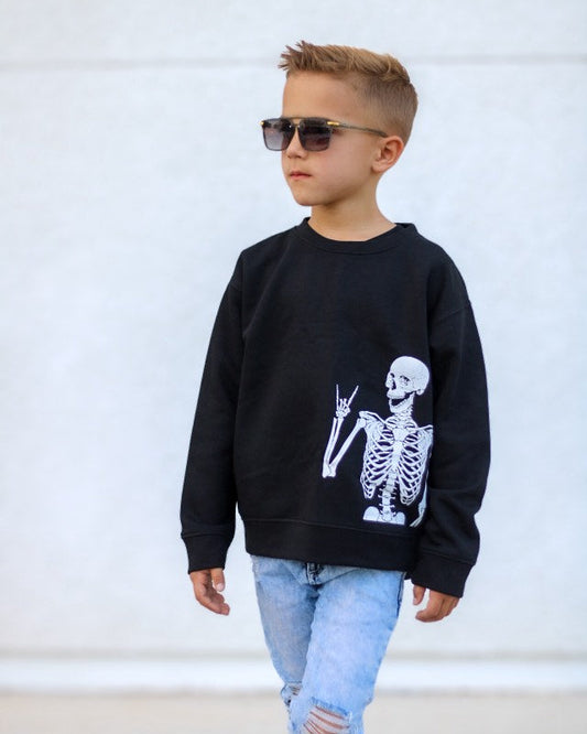 Child wearing a black sweatshirt with a skeleton design, sunglasses, and light blue jeans against a white background.