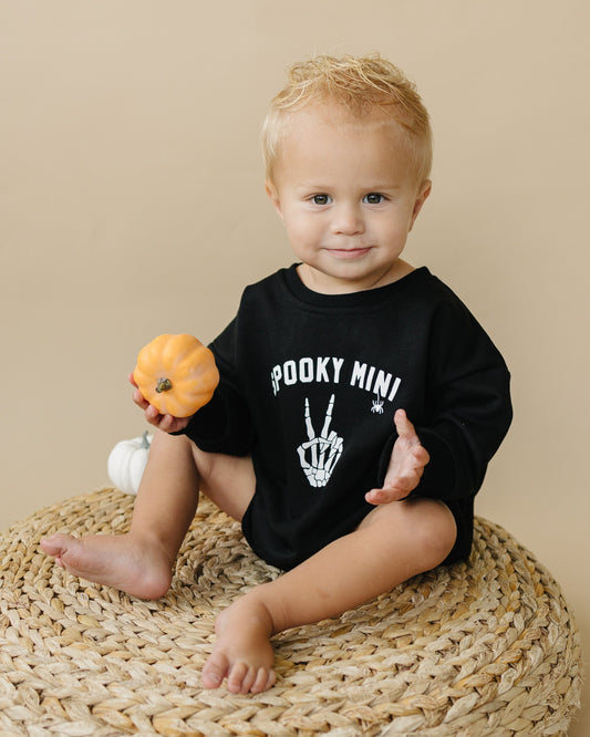 Child wearing a black shirt with 'Spooky Mini' text, holding a small pumpkin on a beige background
