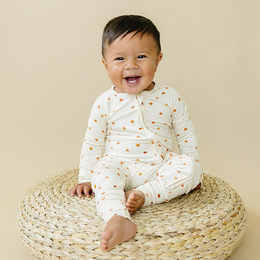 Baby wearing a white onesie with orange patterns, sitting on a woven mat against a beige background