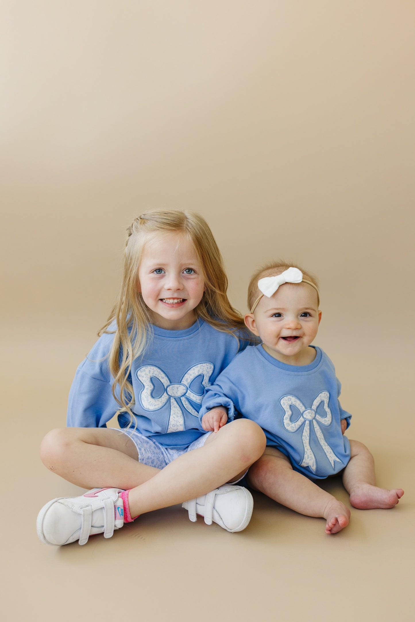 Two young girls wearing matching blue sweatshirts with white bow designs on a beige background.