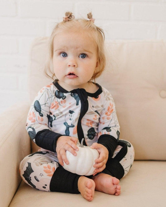 Child wearing a floral onesie with black accents, sitting on a beige couch holding a small white pumpkin.