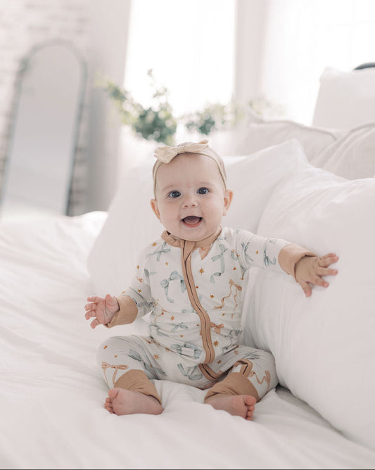 Baby sitting on a bed wearing a patterned onesie in a bright room.