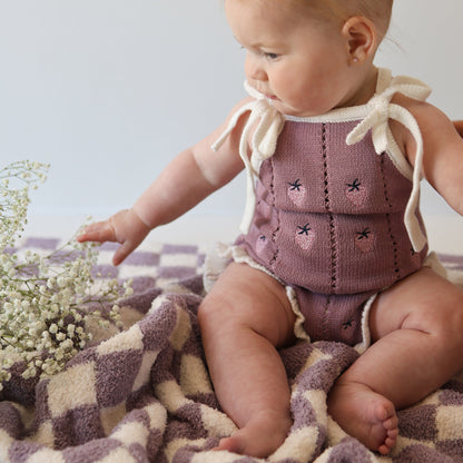 Baby in a pink knitted outfit sitting on a checkered blanket with flowers.