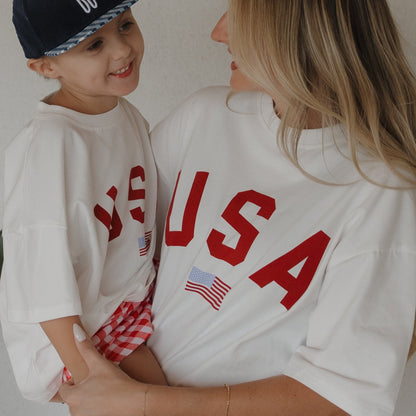 Woman and child wearing 'USA' shirts with an American flag, smiling and looking at each other.
